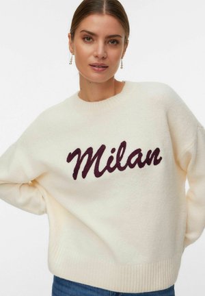 Joven mujer vistiendo un suéter crema con "Milán" en un guion oscuro, posando con una mano en la cadera frente a un fondo blanco liso.