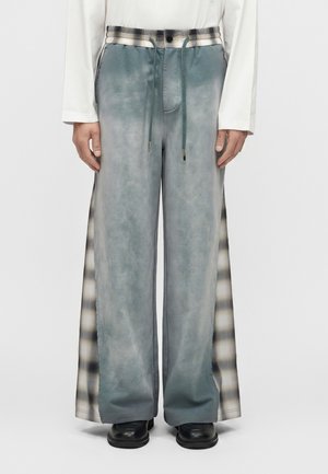 Spodnie o szerokich nogawkach z wyblakłymi niebieskimi panelami z przodu i czarno-białymi kratowanymi panelami po bokach, noszone z czarnymi butami i białą bluzką z długim rękawem.