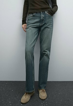 Personne portant un jean bleu délavé à jambes droites, un haut marron côtelé à manches longues et des chaussures beiges en daim à lacets, debout contre un mur gris.