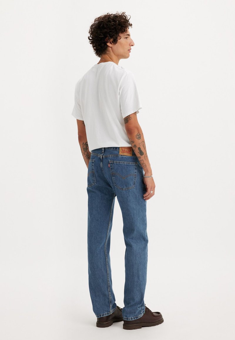 Chemise blanche à manches courtes associée à un jean bleu. Le jean présente une coupe droite et une pièce en cuir sur la taille, avec des détails de couture visibles.
