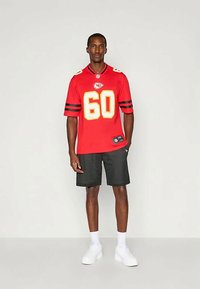 Camisa desportiva vermelha de mangas curtas, apresenta um grande número 60 amarelo e o logótipo da equipa. Combinada com calções de malha pretos e ténis brancos.