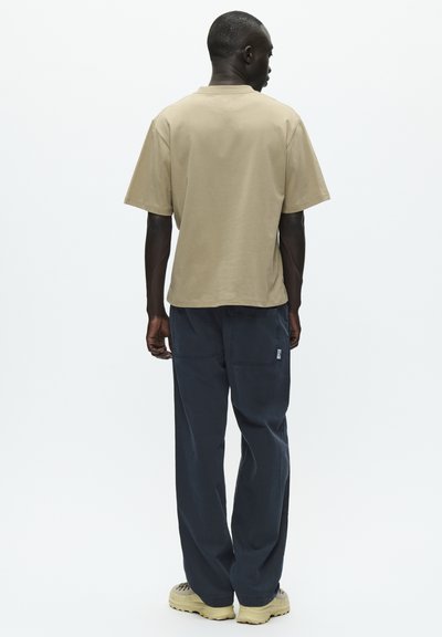 T-shirt en coton beige à manches courtes et à coupe décontractée, associé à un pantalon large bleu foncé avec deux poches arrière et un petit logo.