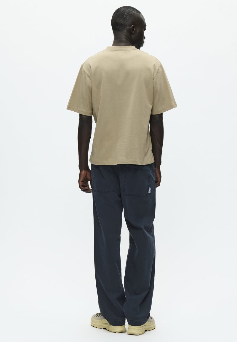 Camiseta de algodón beige con mangas cortas y un corte relajado, combinada con pantalones oscuros holgados de color azul que tienen dos bolsillos traseros y un pequeño logo.
