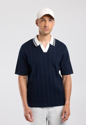 Man draagt een navyblauw, kort mouw gebreid shirt met witte kraag en een lichtgekleurde baseballpet, staand tegen een effen achtergrond.