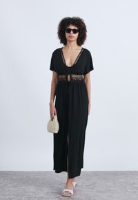 Μαύρο μάξι φόρεμα με κοντά μανίκια, σχέδιο δαντέλας στη μέση και μπροστινή σχισμή. Το μοντέλο κρατάει ένα υφαντό τσάντα και φοράει γυαλιά ηλίου, σε συνδυασμό με ανοιχτόχρωμες σαγιονάρες.