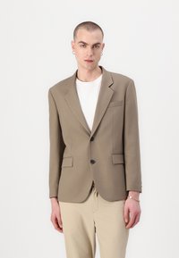 Brown tailored blazer with two front pockets and black buttons. Worn over a white shirt, paired with light-colored pants. Smooth texture.