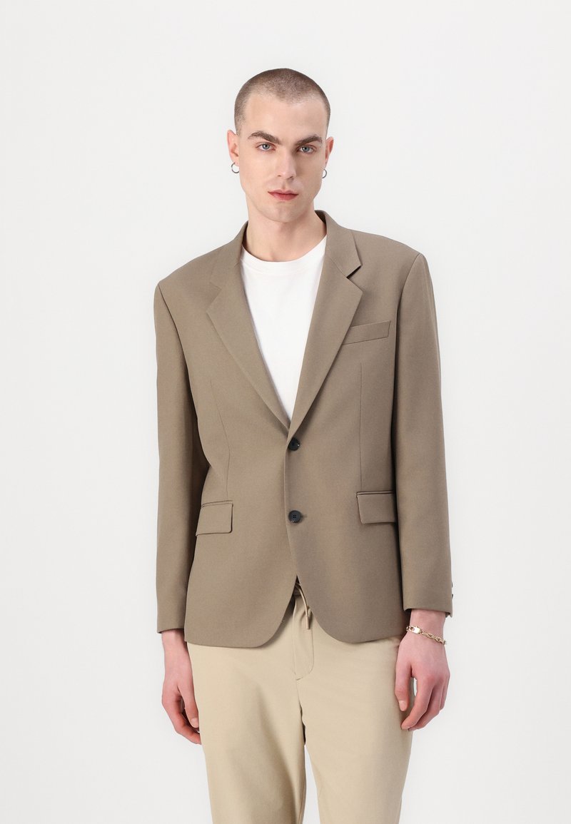 Brown tailored blazer with two front pockets and black buttons. Worn over a white shirt, paired with light-colored pants. Smooth texture.