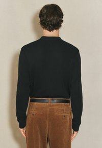 Schwarz langärmliges Shirt mit strukturiertem Ribbel, kombiniert mit hellbraunen Cordhosen und einem schwarzen Ledergürtel. Rückansicht.