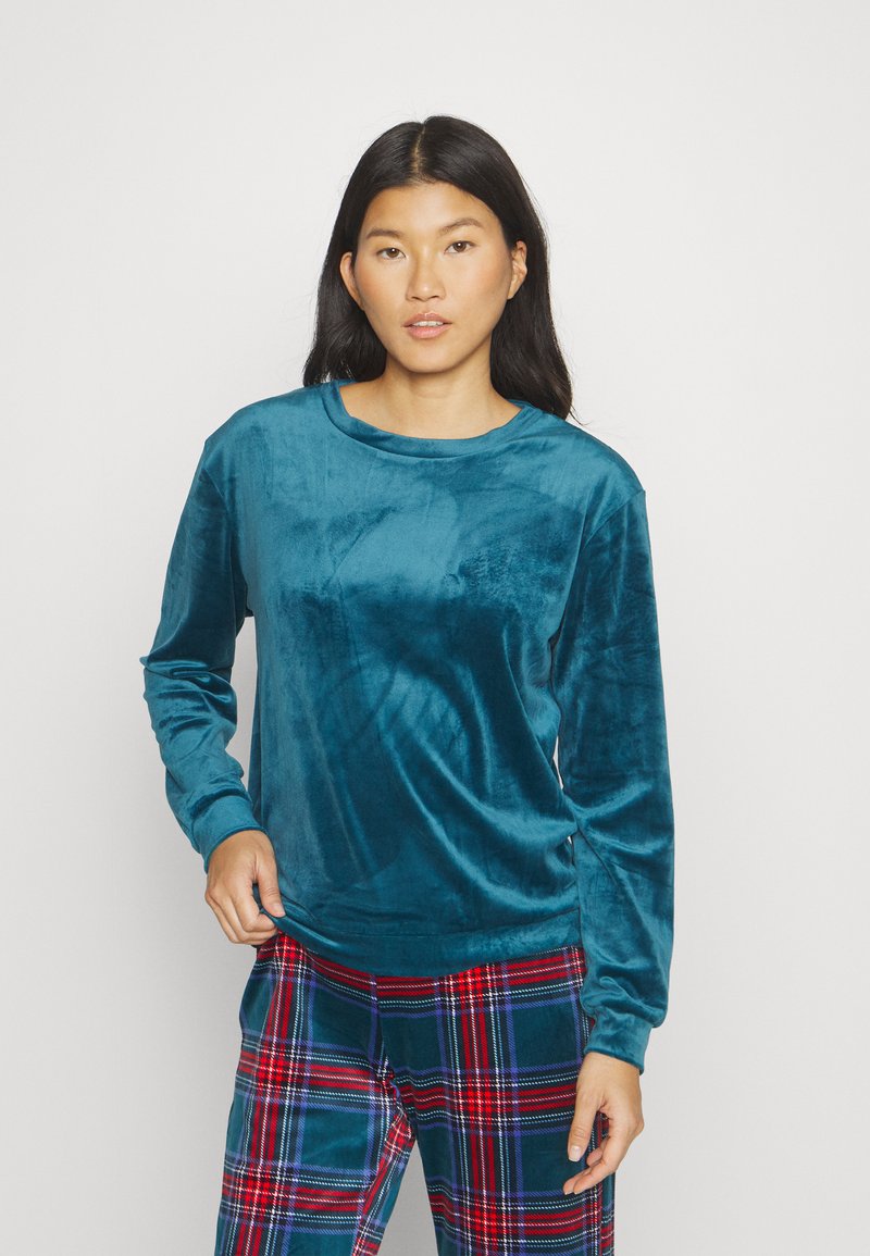 Haut en velours teal à manches longues avec un col ras du cou, texture lisse et coupe décontractée, associé à un pantalon à carreaux avec des tons rouges et verts.