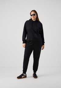 Felpa nera con cappuccio e pantaloni della tuta coordinati realizzati in tessuto morbido. Il modello indossa occhiali da sole con montatura bianca e scarpe da ginnastica nere con suola marrone.