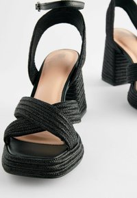 Černé wedge sandály s tkaným svrchním designem, se zkříženými pásky a čtveratou špičkou. Hladká vnitřní podšívka a detail popruhu kolem kotníku.
