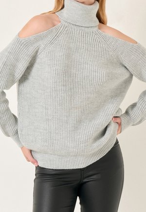 Femme portant un pull à col roulé gris clair côtelé avec épaules dénudées et un pantalon en cuir noir ajusté, les mains sur les hanches.