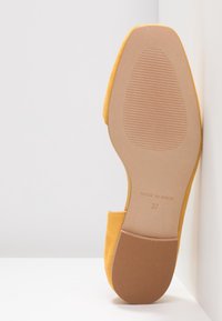 Chaussure plate jaune appuyée contre un mur blanc, montrant la semelle texturée marquée "Fabriqué en Espagne" et la taille 37.