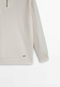 Pullover beige a maniche lunghe con collo a mezza zip, polsini e orlo a costine, con un piccolo etichetta nera vicino al fondo.