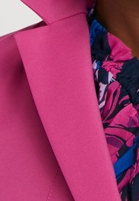 Jasně růžové sako přes tmavý květinový top s volánkovým výstřihem na osobě se středním tónem pleti.