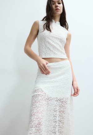 Donna con top corto senza maniche in pizzo bianco e gonna a vita alta in pizzo abbinata con la metà inferiore trasparente su sfondo chiaro semplice.