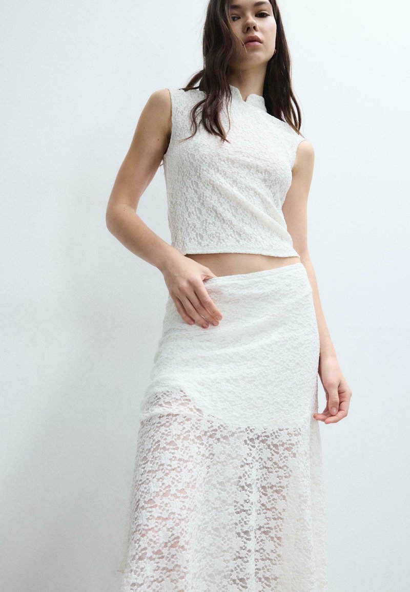Frau in weißem ärmellosem Spitzen-Crop-Top und passendem hoch tailliertem Spitzenrock mit transparentem unterem Teil vor schlichtem, hellem Hintergrund.