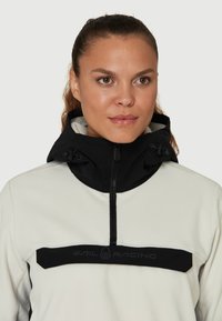 Polarowy półgolf w białym kolorze z czarnym kapturem i panelem na piersi. Posiada zamek błyskawiczny z przodu oraz akcent logo. Gładka tekstura i sportowy design.