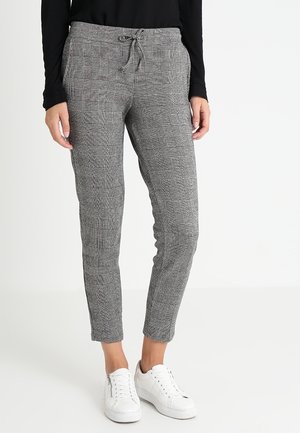 Kobieta w szarych kraciastych spodniach z troczkami i kieszeniami oraz białych tenisówkach, stojąca na tle jednolitego jasnego tła.