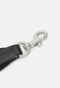 Mousqueton pivotant en métal argenté attaché à une lanière en cuir noir avec des coutures visibles sur un fond clair uni.