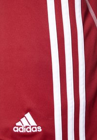 Punainen urheilupaita, jossa on valkoiset raidat sivuilla. Paidassa on pieni valkoinen Adidas-logo lähimpänä alareunaa. Sujuva ja kevyt kangas.