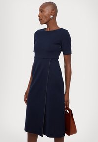 Tengerészkék, rövid ujjú ruha, which fitted bodice, bővülő szoknyával és cipzáros részletekkel. Barna kézitáska az egyik kézben. Sima textúra.