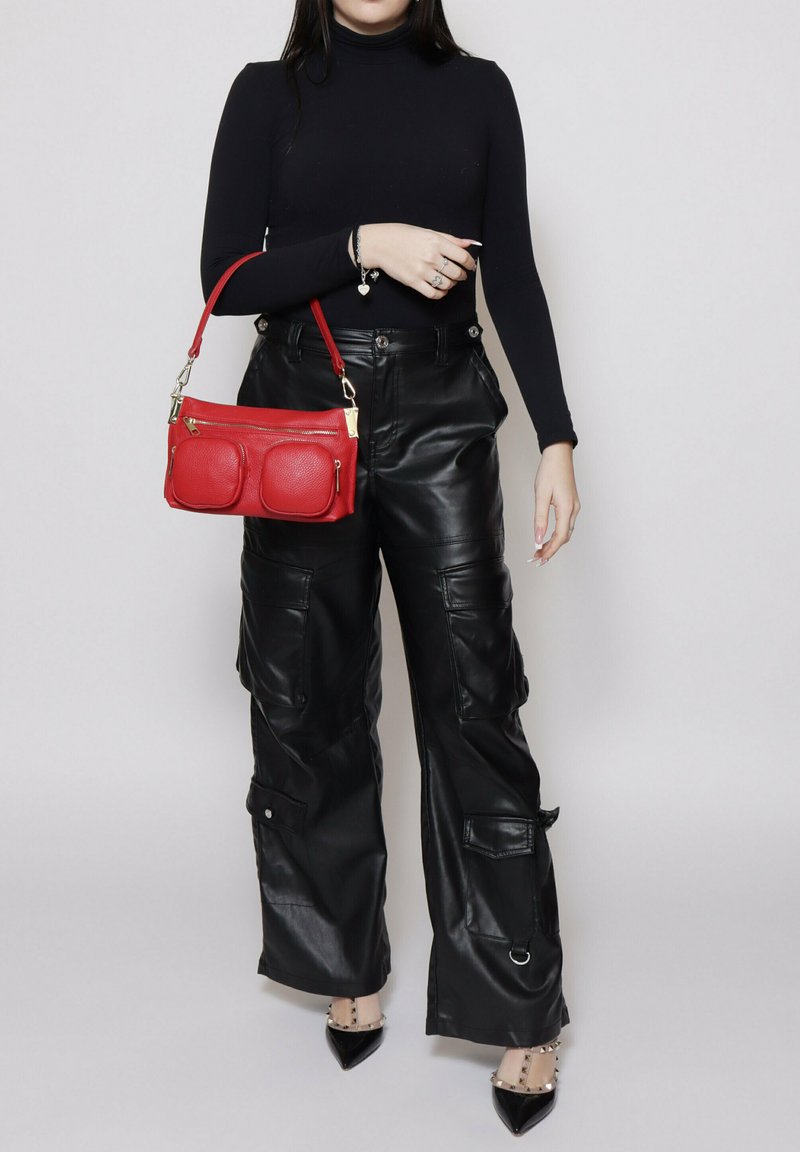 Donna con dolcevita nero e pantaloni cargo in pelle che tiene una borsa rossa con dettagli dorati, in piedi davanti a uno sfondo semplice.