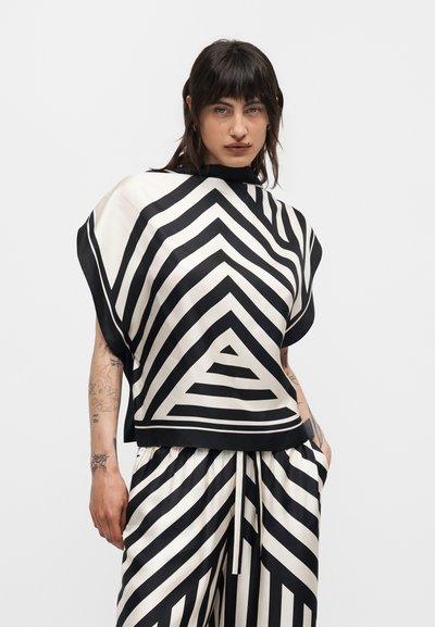 Άτομο με σκούρα μαλλιά που φοράει ένα μαύρο και λευκό γεωμετρικό ριγέ τοπ και ταιριαστό παντελόνι, στέκεται μπροστά σε ένα απλό φωτεινό φόντο.