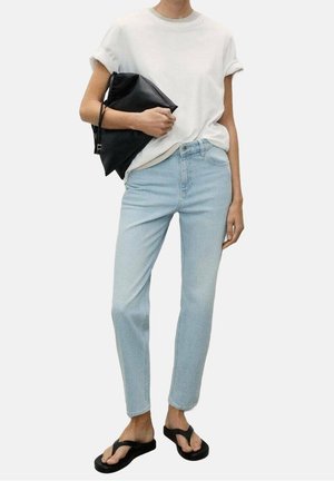 Person trägt hellblaue Jeans, weißes T-Shirt mit hochgekrempelten Ärmeln, schwarze Sandalen und hält eine große schwarze Clutch unter einem Arm.
