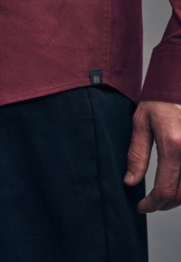 Bordo majica dugih rukava s teksturiranim završetkom i malom oznakom branda na rubu, uparena s tamnim hlačama, prikazuje detaljan prikaz ruke.