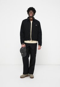 Chaqueta negra con cremallera, suéter beige, pantalones negros, gorra gris, botas marrones. Presenta un pequeño logo en la chaqueta, corte relajado y detalles de costura visibles.