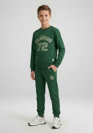Tienerjongen staat en lacht, draagt een groene trui en joggingbroek met de tekst "Champions 72", en witte sneakers op een effen achtergrond.