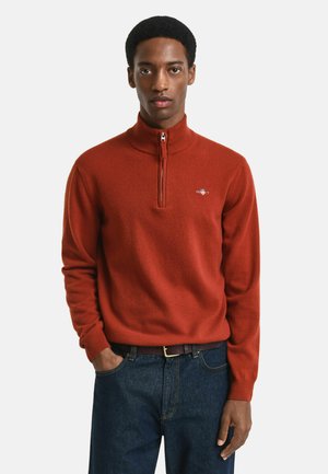 Pullover rosso in fleece con colletto alto e design con mezzo zip. Presenta polsini a coste e un piccolo logo sul petto. Abbinato a jeans scuri.