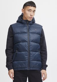 Mörkblå pufferväst med huva, dragkedja fram och quiltad struktur; har sidofickor och svarta ärmar. Logotyp på bröstet.