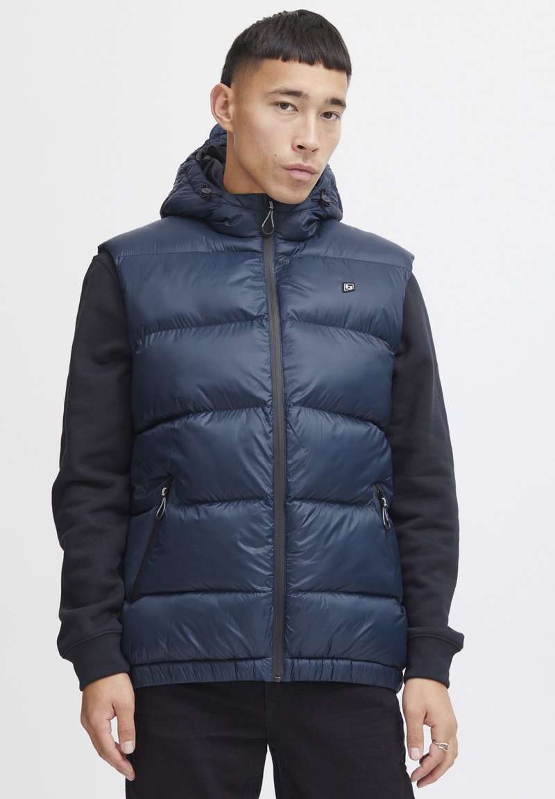 Mörkblå pufferväst med huva, dragkedja fram och quiltad struktur; har sidofickor och svarta ärmar. Logotyp på bröstet.