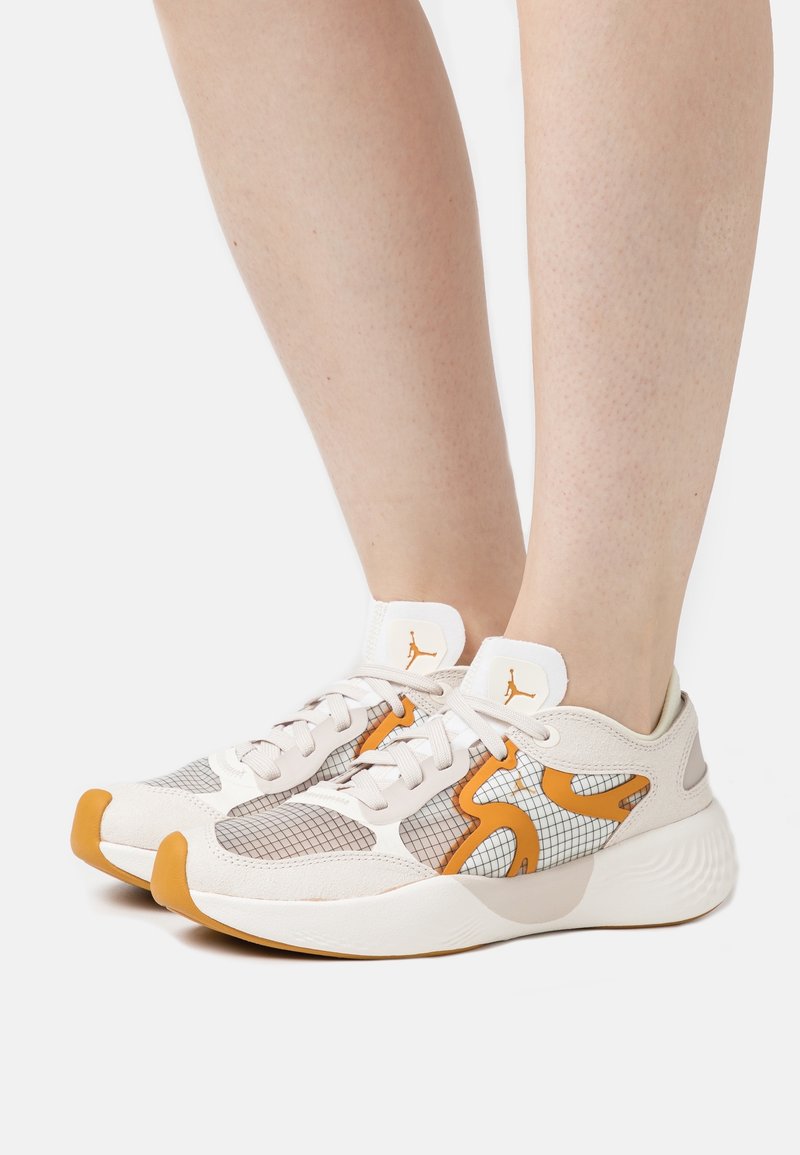 Chaussures de sport beige clair et orange avec lacets blancs portées sur jambes nues, présentant un motif quadrillé et des semelles rembourrées.