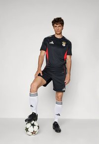 Črno nogometno oblačilo z rdečimi poudarki, s logotipom Adidas, črtastimi belimi nogavicami in črnimi kopačkami. Igralec stoji na nogometni žogi.