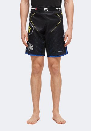 Homme portant un short de combat Venum Tekken 8 noir et bleu avec des logos, debout pieds nus sur un fond uni.