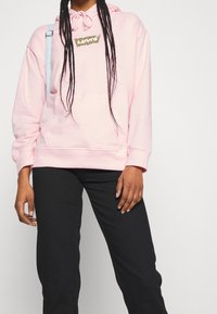 Hoodie rosa realizzato in misto cotone, con tasca frontale e logo a stampa leopardata. Abbinato a pantaloni neri per un contrasto.