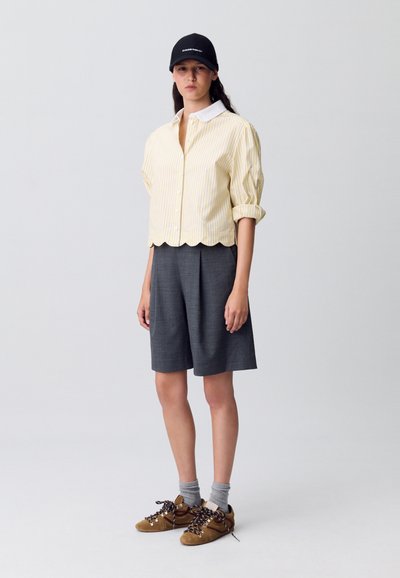 Camicia a righe gialla con colletto bianco, pantaloni corti grigi plissettati, calze grigie e scarpe marroni con lacci. Design semplice e casual.