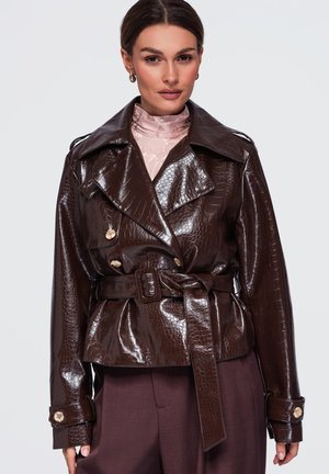 Femme portant une veste en similicuir marron foncé brillante ceinturée avec un motif texturé, sur un haut rose clair à col montant et un pantalon foncé.
