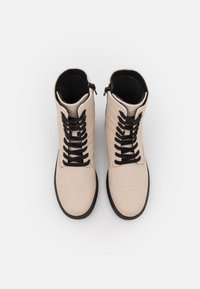 Botas de piel beige hasta el tobillo con cordones negros y cremallera lateral, con una textura suave y punta redondeada, sobre un fondo blanco.