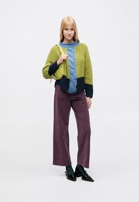 Adīts džemperis zaļā, tumši zilā un zilā krāsā ar pintes rakstu; saskaņots ar plum krāsas platām biksēm un melniem apaviem. Modelis tur nelielu zaļu somiņu.