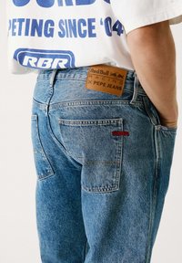 Jeans de mezclilla azul claro con un parche de cuero que dice "Red Bull Racing x Pepe Jeans" y el logo rojo "RBR" en el bolsillo trasero.