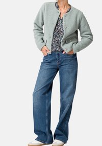 Lichtgroene, gestructureerde fleecjas met een ronde hals en zilveren knopen, gecombineerd met wijde blauwe jeans en witte sneakers.