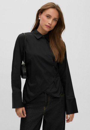 Femme aux longs cheveux châtain clair portant un chemisier noir asymétrique boutonné, un jean noir, des boucles d'oreilles en perles et un petit sac noir porté à l'épaule.