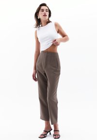 Witte mouwloze top gecombineerd met hoogwaardig, geplooide bruine broek, met zijzakken en een getailleerde pasvorm, gedragen met zwarte sleehak sandalen.