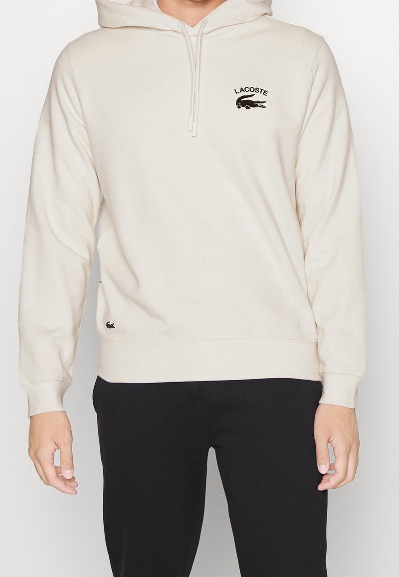 Personne portant un sweat à capuche Lacoste beige clair avec cordons et un pantalon noir, debout devant un fond blanc uni.