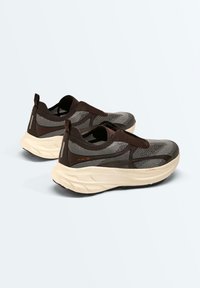 Een paar donkergrijze en zwarte slip-on sportieve sneakers met dikke witte zolen en bovenzijdes van gaasstof op een lichte achtergrond.