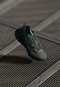 Scarpa sportiva verde con superficie texturizzata, design dei lacci intricato e suola in gomma prominente, sospesa sopra una griglia metallica a motivi.